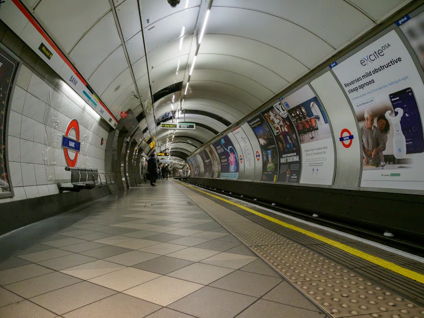 An underground metro station platform illuminated by artificial lighting, with a curved ceiling and tiled walls decorated with advertisements featuring urban scenes and brands. The platform is empty, 
